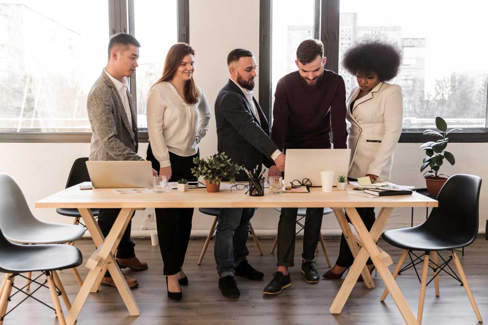 Team of business professionals standing around a desk reviewing a laptop and discussing strategy, representing collaborative planning and expert transaction support through M&A Advisory New Jersey services.